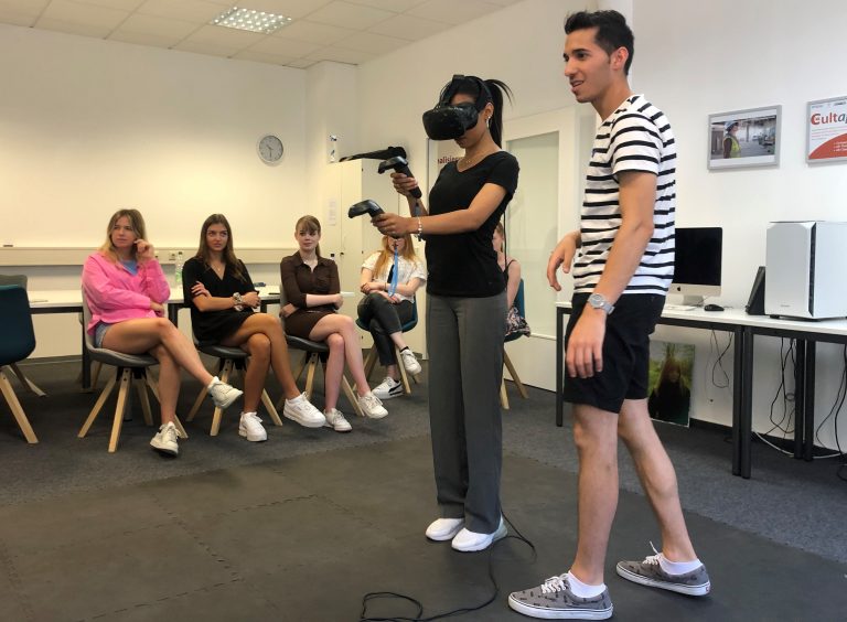 FHM-Medienpsychologie-Studentin Joyce- Madison Etienne teste das VR-Gerät im hauseigenen VR-Labor. Eyad Sattout (FHM) erklärt ihr die Technik. Im Hintergrund v.l. Sophie Borchard, Marie Joline Sendker, Nina Kruggel und weitere Studentinnen der Studiegruppe. (Foto: Fachhochschule des Mittelstands (FHM) )