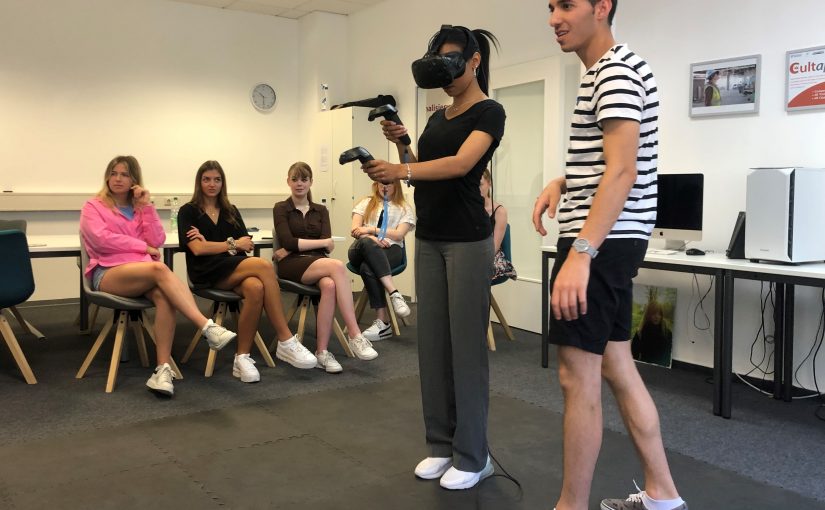 FHM-Medienpsychologie-Studentin Joyce- Madison Etienne teste das VR-Gerät im hauseigenen VR-Labor. Eyad Sattout (FHM) erklärt ihr die Technik. Im Hintergrund v.l. Sophie Borchard, Marie Joline Sendker, Nina Kruggel und weitere Studentinnen der Studiegruppe. (Foto: Fachhochschule des Mittelstands (FHM) )