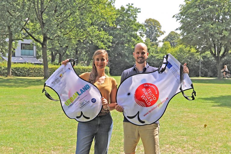 Melanie Adriaans, REGIONALE 2022, und Markus Backes, Teutoburger Wald Tourismus, stellen den Wickelfisch vor. Die OstWestfalenLippe GmbH unterstützt das 1. Mindener Weserschwimmen der DLRG mit 500 kostenfreien Wickelfischen. (Foto:OstWestfalenLippe GmbH)