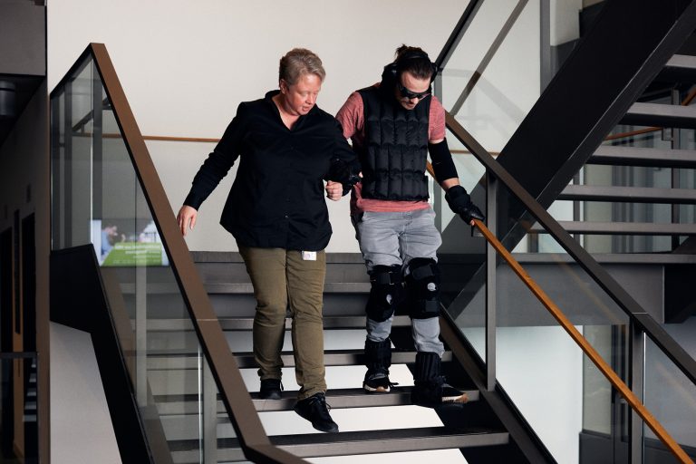 Ariane Rolf arbeitet im Skills Lab an der HSBI. Sie hilft Student Jerome Sievers im Alterssimulationsanzug die Treppe herunter. (Foto: P. Pollmeier/HSBI)