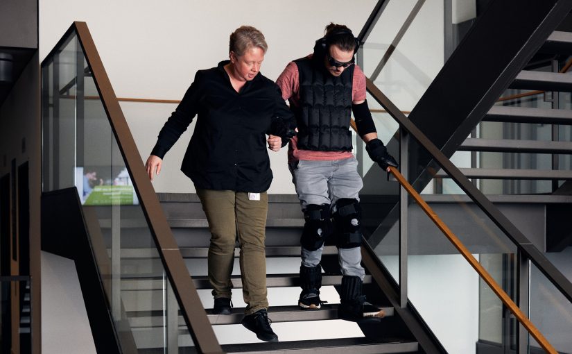 Ariane Rolf arbeitet im Skills Lab an der HSBI. Sie hilft Student Jerome Sievers im Alterssimulationsanzug die Treppe herunter. (Foto: P. Pollmeier/HSBI)