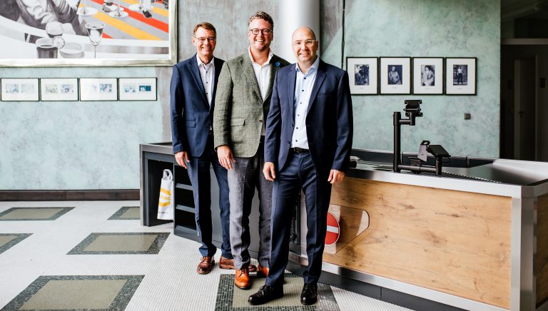 Peter Weichert, Geschäftsführer HARTING Systems, Philip Harting, Vorstandsvorsitzender der HARTING Technologiegruppe und Dr. Christoph Barth, designierter Geschäftsführer der neuen HARTING Systems GmbH (v. l.) vor einer der Kassenzonen, die HARTING Systems produziert hat. (Foto: Harting)
