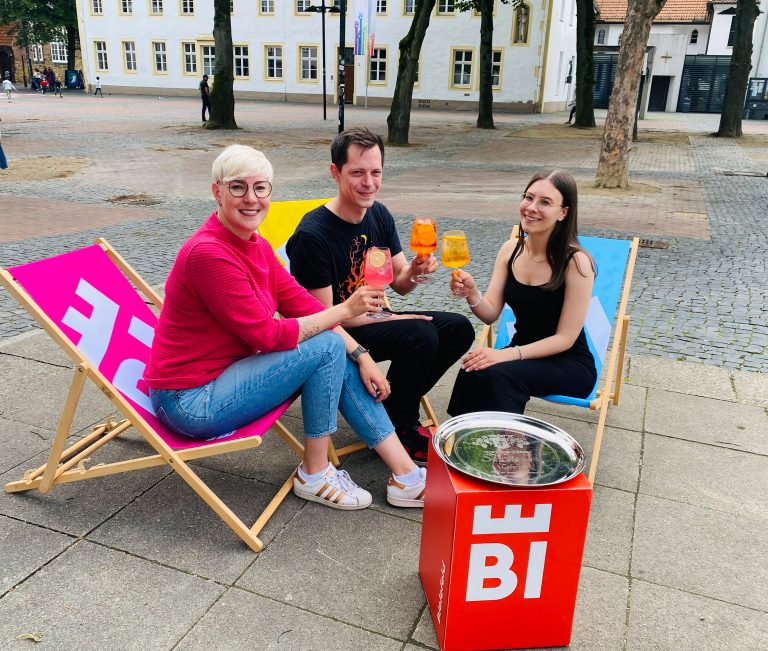 Katharina Schilberg, Jens Kramer und Leonie Klau (v. l.) von Bielefeld Marketing sind bereit für die Abendmarkt-Saison 2024. Jeden Donnerstag, vom 4. Juli bis 22. August, gibt es auf dem Klosterplatz Gastro-Stände und Live-Musik. (Foto: Bielefeld Marketing GmbH)