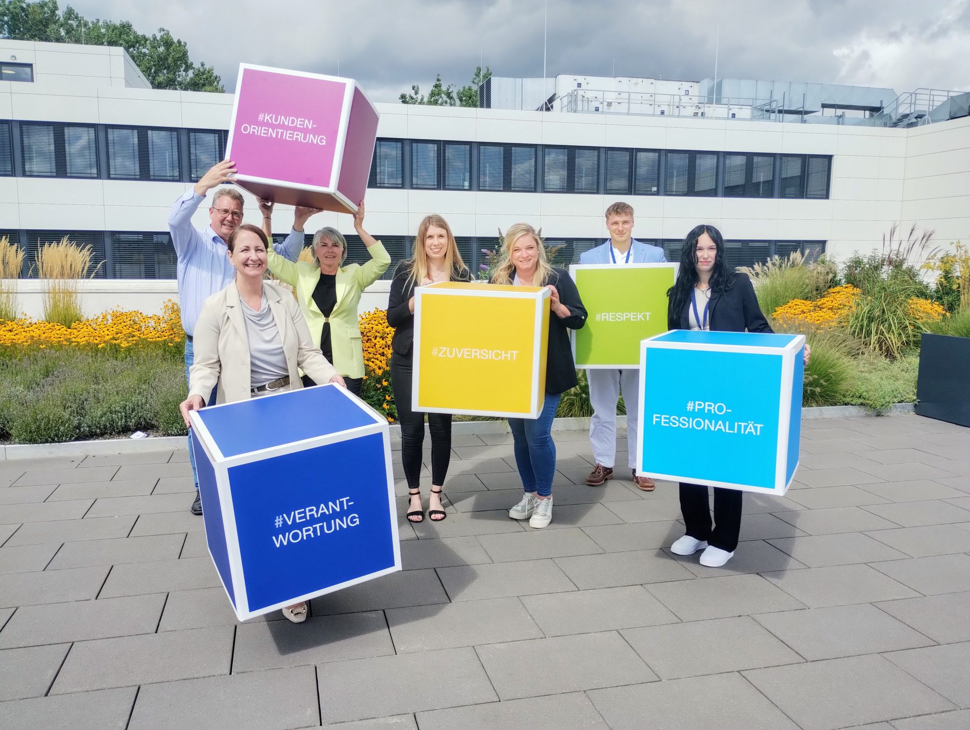 Unternehmenswerte kreativ präsentiert: Birgit Aspin (Chief People Officer und Mitglied der Geschäftsleitung) sowie (v. l.) Frank Schultz (Abteilungsleiter Direktservice und Ausbildungswerkstatt), Nadja Bürger (Leiterin Geschäftsfeld Ambulantes Gesundheitswesen, Direktservice und Ausbildungswerkstatt und Mitglied der Geschäftsleitung) und Leonie Münnich (Direktservice-Team / Ausbildungswerkstatt), Pia Stohlmann (Referentin Personalentwicklung) begrüßen mit Niklas Kehne (Auszubildender zum Kaufmann für Versicherungen und Finanzen) und Lilli Richter (Auszubildende zur Kauffrau für Versicherungen und Finanzen) zwei der neuen Auszubildenden. (Foto: Ecclesia Gruppe)
