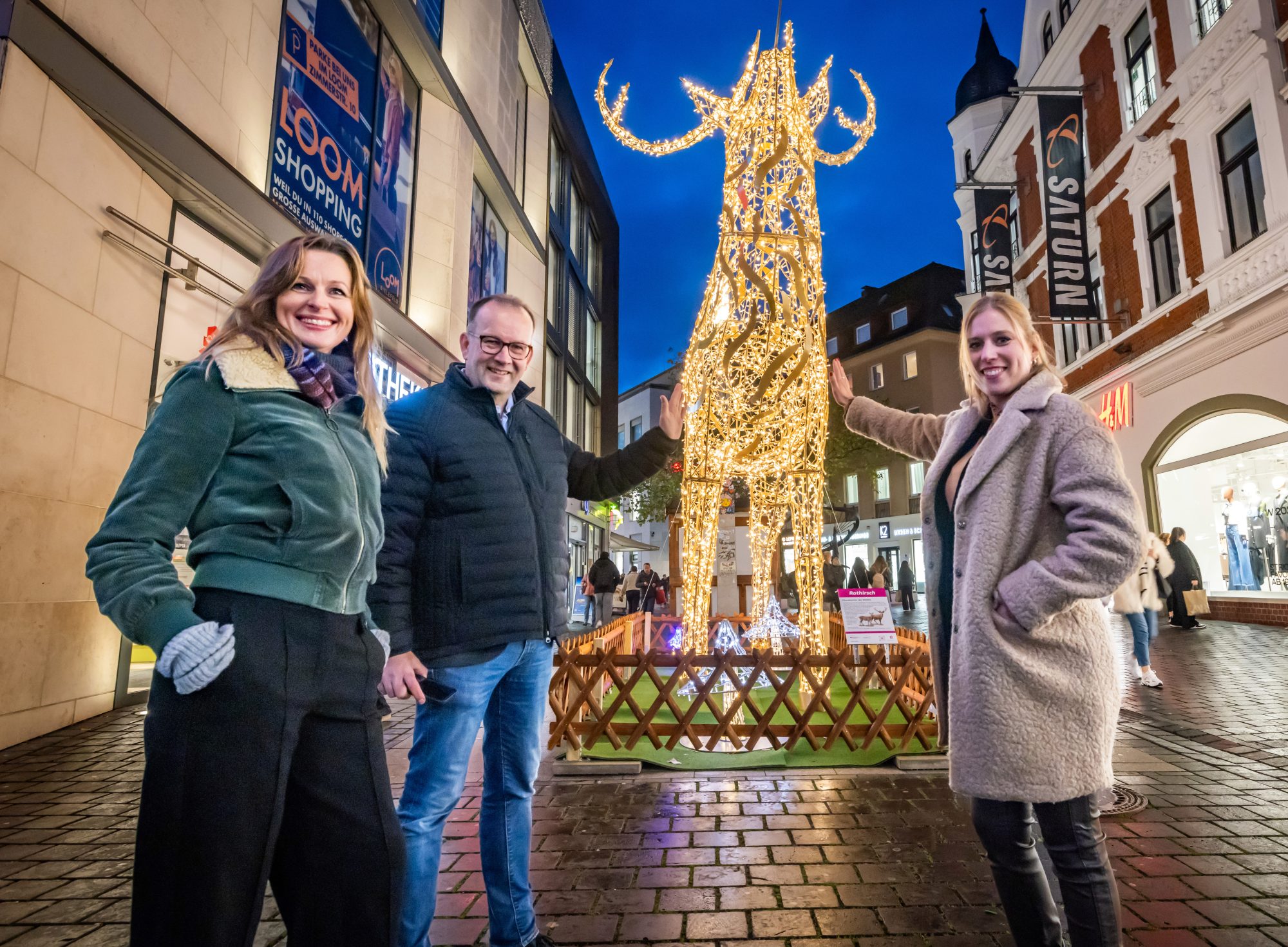 Birgit Schroers, Martin Knabenreich und Julia Lehmann (v. l.) von Bielefeld Marketing präsentieren das Lichtprojekt „Von hier wech“ am leuchtenden Hirsch an der Bahnhofstraße. (Foto: Bielefeld Marketing | Sarah Jonek)