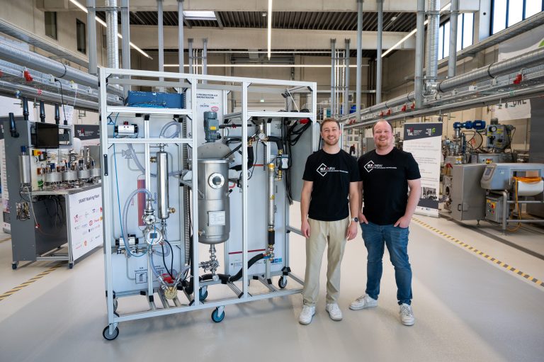 Athur Gossen (l.) und Marc Trilling-Haasler vor ihrer Hafermilch-Anlage. Diese soll ein Beispiel dafür sein, wie ein transparenter Lebensmittelproduktionsprozess in Zukunft aussehen kann, und welche Vorteile er sowohl für Unternehmen als auch für Verbraucher:innen bietet (Foto: TH OWL)