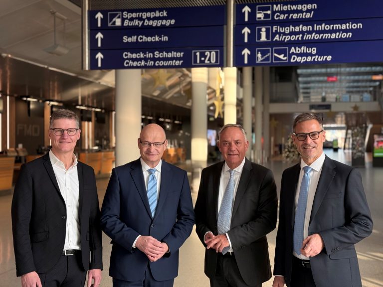 Vier Personen im Terminal des Flughafens Münster/Osnabrück vor Hinweisschildern – Führungswechsel am FMO
