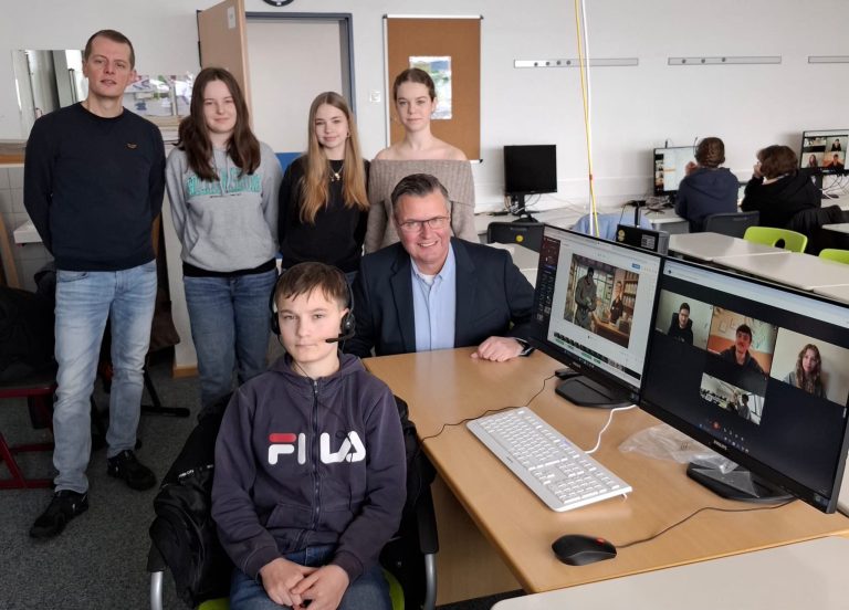 Schülerinnen und Schüler präsentieren ein KI-generiertes Video im Klassenraum; auf Monitoren sind Videokonferenz und Filmszene zu sehen