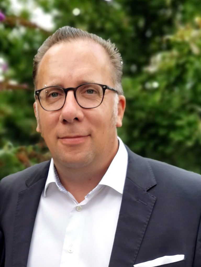 Porträt von Thomas Werning im Sakko und weißem Hemd vor unscharfem Grün im Hintergrund