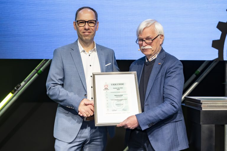 Zwei Männer bei der Urkundenübergabe auf einer Bühne, einer hält eine gerahmte Auszeichnung für fetra.