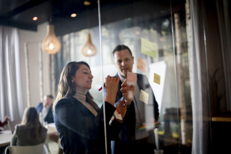 Zwei Berufstätige arbeiten in einem Besprechungsraum mit Haftnotizen an einer Glasscheibe und diskutieren über Themen der Zusammenarbeit.