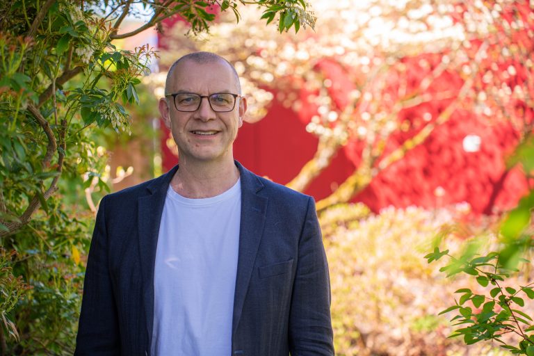 Dr. Christoph Harrach steht im Außenbereich des Campus Höxter vor grünen Pflanzen und einem roten Hintergrund.