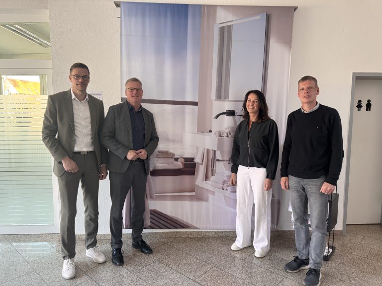Vier Personen stehen bei einem Unternehmensbesuch der Stadt Rheine und der EWG Rheine in den Räumen der Dyckhoff GmbH vor einer großformatigen Textilaufnahme.