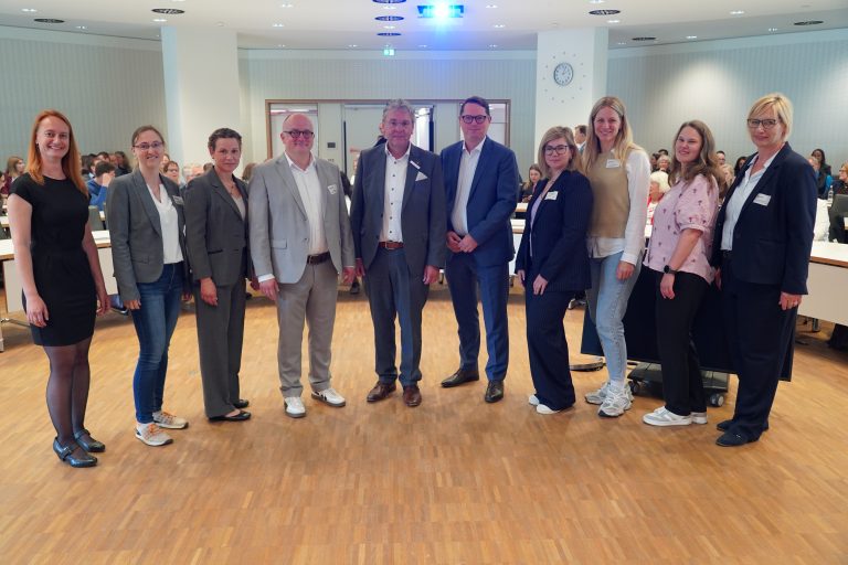 Gruppenbild von neun Personen bei einer Fachtagung in einem voll besetzten Veranstaltungsraum