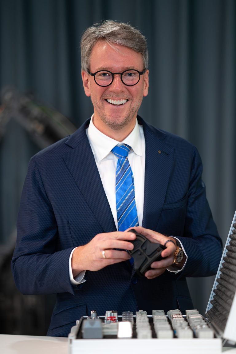 Philip Harting hält ein technisches Bauteil in der Hand und steht vor einem Musterkoffer mit industriellen Komponenten