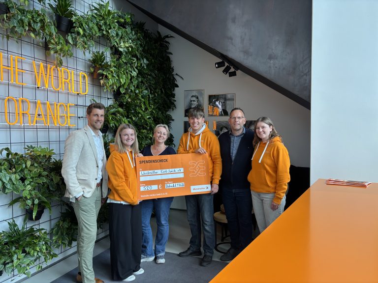 Sechs Beteiligte stehen in der Weidmüller Welt in Detmold und halten einen orangefarbenen Spendenscheck über 520 Euro.