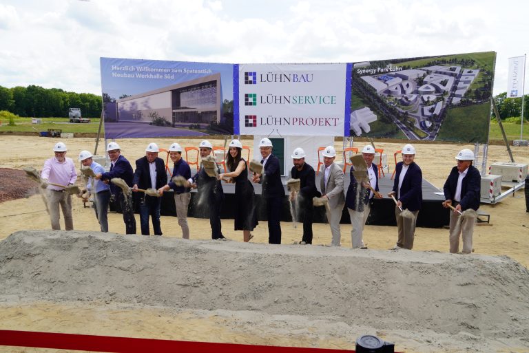 Beteiligte beim ersten Spatenstich für den Synergypark Lühn in Lingen vor einer Baustellenbühne mit Projektvisualisierungen